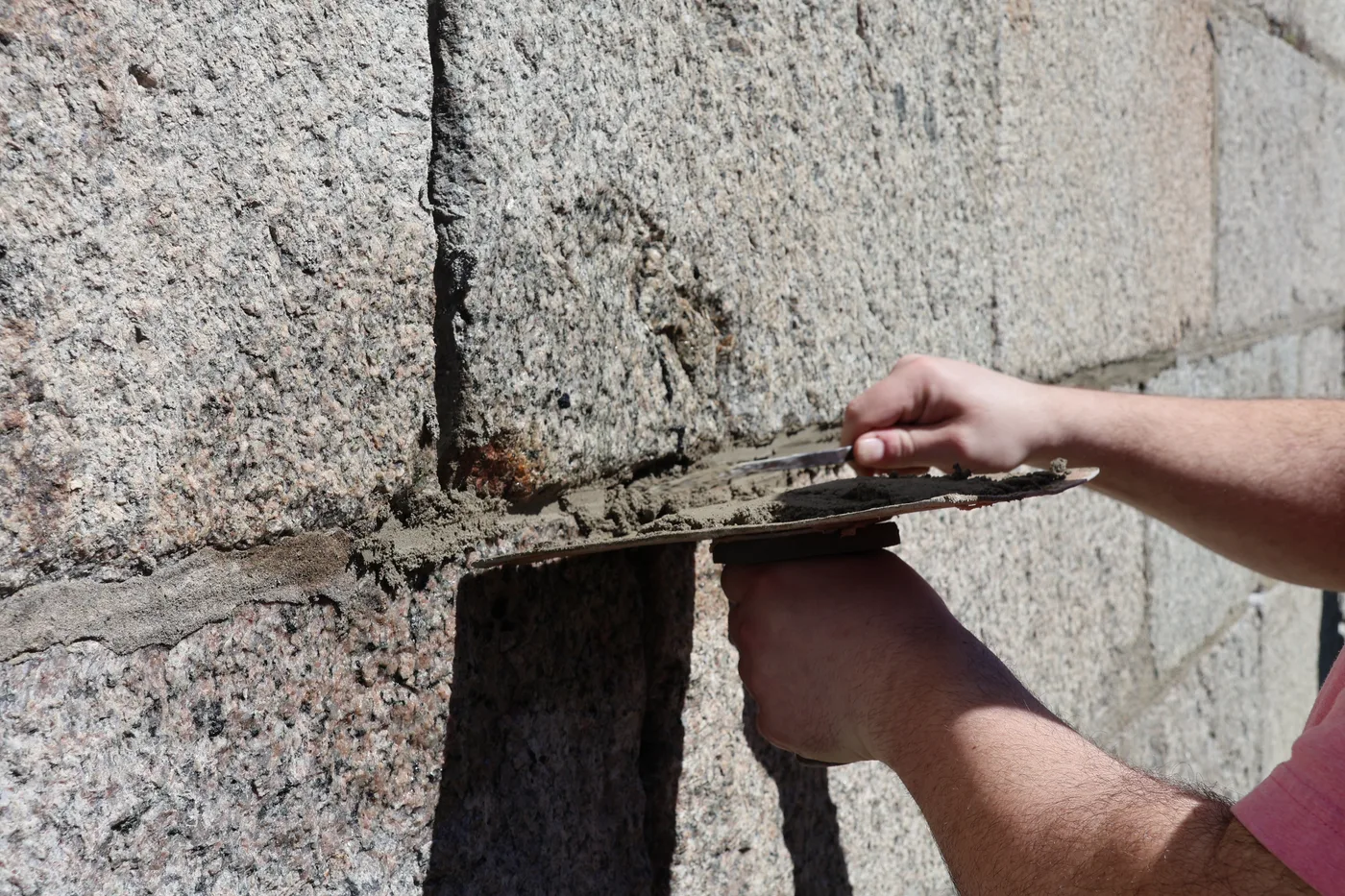Hands applying mortar between granite blocks in a Fort Adams wall